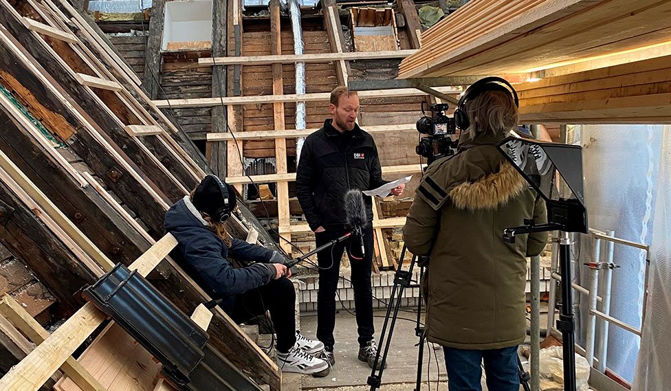 En person står og snakker ind i kamera oppe på et tag der bliver renoveret.