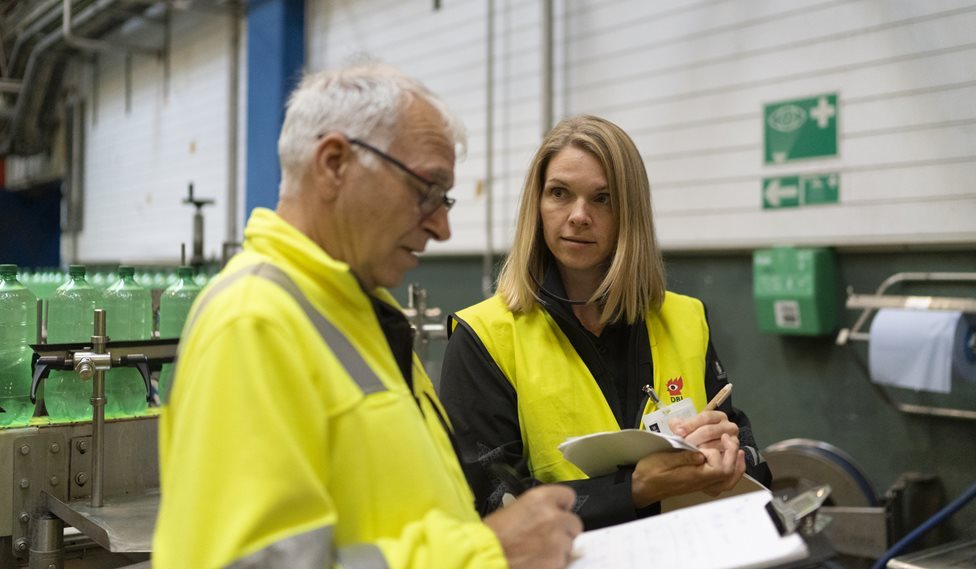 To personer står i en fabrik og smakker og den ene kigger ned i sine papirer